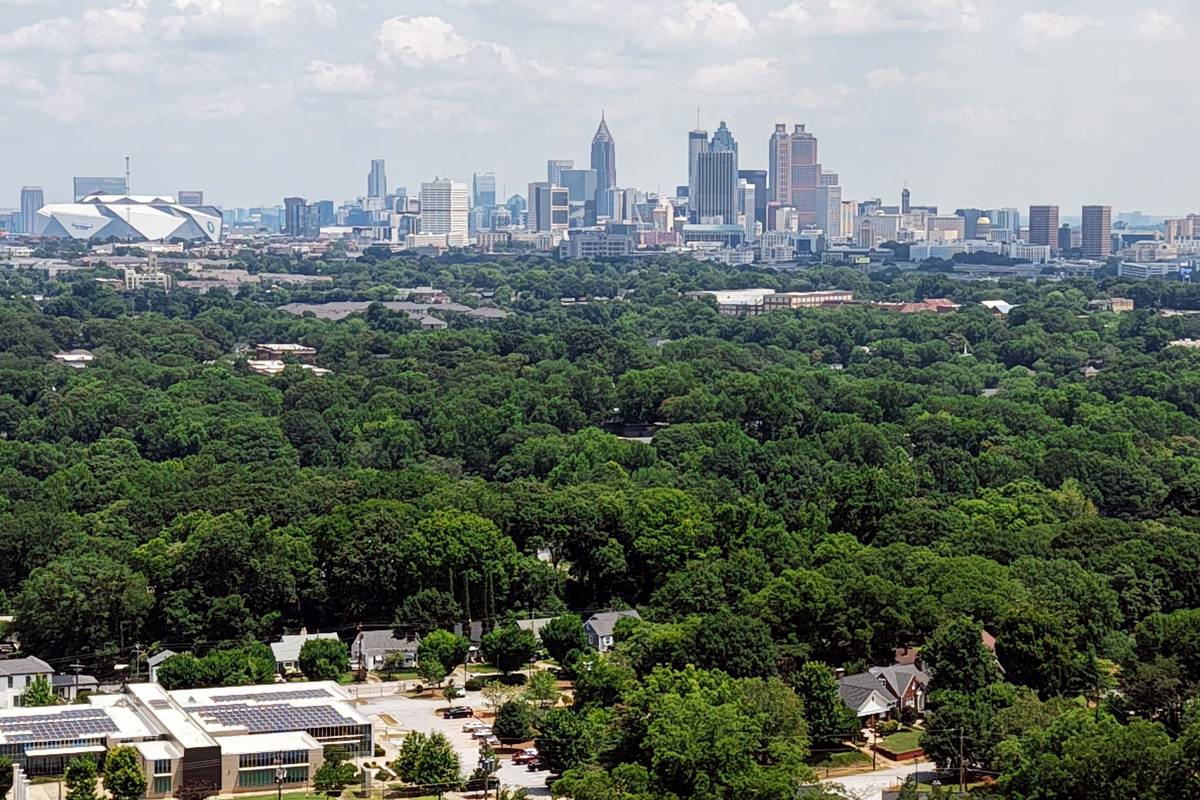 Aerial view across dense green tree canopy toward the downtown Atlanta skyline, with a stadium and low residential blocks in the foreground.